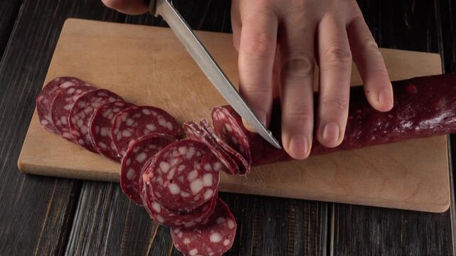 Hands slicing sausage into thin rounds on a wooden cutting board, showcasing the continuous action of food preparation in a kitchen setting