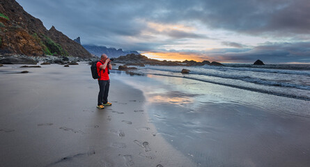 A male photographer wearing a bright orange t-shirt stands on the wet volcanic black sand of Playa de Benijo while photographing the sunset over the Atlantic © Xalanx