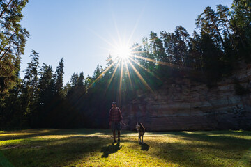 A traveler in a plaid shirt and his husky dog stand in a sunny forest clearing against a majestic cliff. © Dmitri