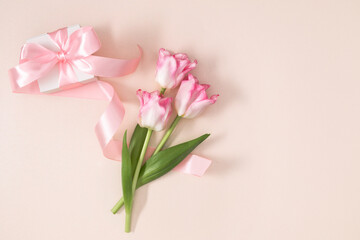 Obraz premium Pink tulip flowers and white gift box with silk ribbon on pastel beige background, top view flat lay with copy space. International Women's Day 8 March greeting card concept. Spring celebration.
