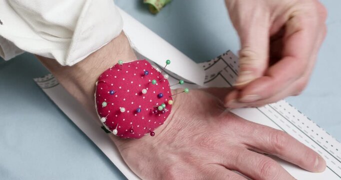 Experienced man tailor chooses sewing pin to securely fix paper pattern onto fabric put across worktable. Professional craftsman attaches pattern to cloth