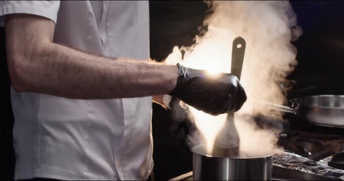 Chef mixes fast dish in stainless saucepan with wooden shovel in kitchen closeup. Master cooks hot food with rising steam in restaurant. Cookware and stove usage