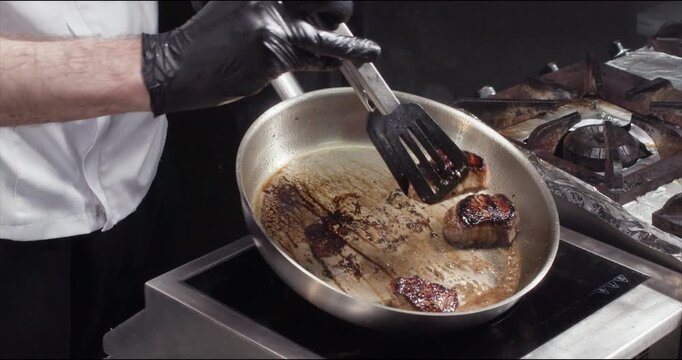 Chef checks meat steaks condition on frying pan with hot oil closeup. Man hands in gloves use tongs to take roasted beef pieces on stove in fancy restaurant