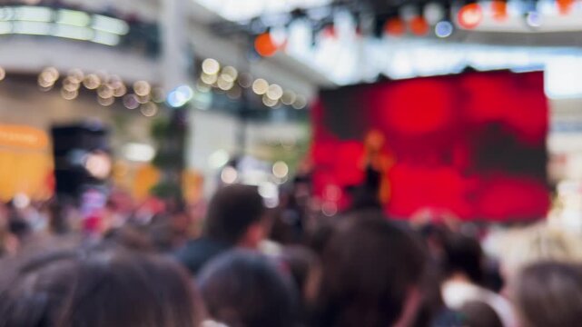 A crowd of young fans waving their hands in the fan zone of a shopping mall atrium. A female singer is performing on stage for an enthusiastic audience gathered for a live event. Out of focus.