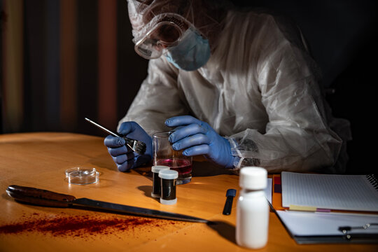 Crime scene evidence processing on wooden desk: gloved hands use a brush to reveal latent prints on a drinking glass beside a knife with blood stains and sample vials.