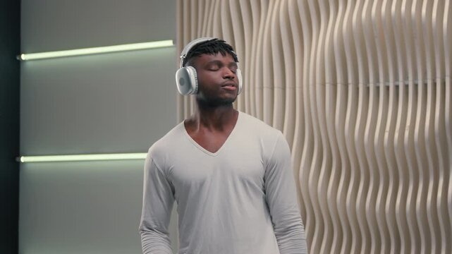 Dancing satisfied African American man in headphones listening music in futuristic room dance alone entertainment with dynamic movements male guy in earphones online playlist in white futurism studio
