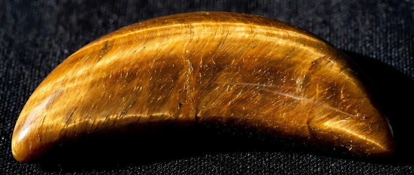 Golden brown tiger eye gemstone cabochon with natural chatoyancy and silk-like luster on dark textured fabric background for jewelry making and lapidary work.