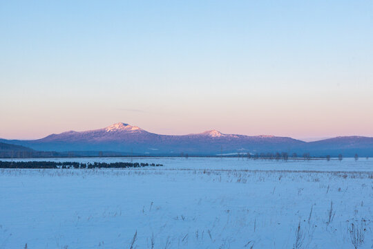 Ural Mountains near the city of Beloretsk, Republic of Bashkortostan. Snow-covered landscape with distant mountains under pastel sky at dawn, serene winter scene in rural area