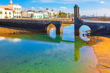 Puente de las Bolas in Arrecife, Lanzarote spans a calm lagoon with volcanic stone arches. Whitewashed buildings, palms and blue skies reflect in water at low tide