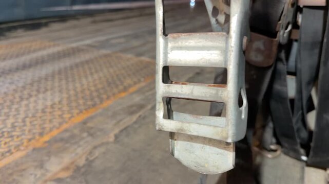 A static close up shot of a metal cargo ratchet and hanging straps inside the bed of a semi trailer as an electric pallet jack drives past in the background