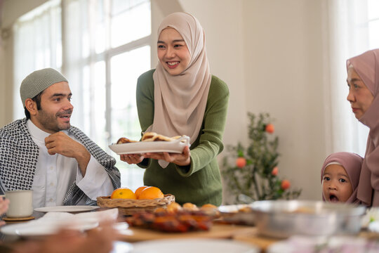 Happy asian muslim woman in hijab serving food plate to family at dining table, arab husband, grandmother and little daughter waiting for meal, Ramadan Iftar, Eid hospitality and halal dining concept