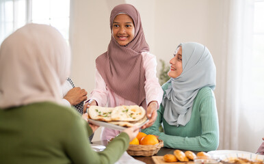 Happy young asian muslim girl in hijab serving flatbread plate to mother at dining table, daughter helping family during meal, good parenting, ramadan Iftar, teaching values and halal food concept