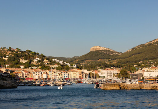 Entr&eacute;e du port du village de Cassis en &eacute;t&eacute;, vue depuis la mer, bateaux, maisons de village, collines en arri&egrave;re-plan, ciel bleu, soleil de fin d&rsquo;apr&egrave;s-midi, vie douce en M&eacute;diterran&eacute;e, Sud de la France