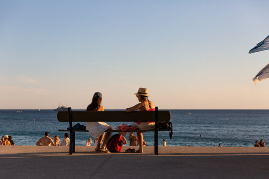 Femmes vue de dos face &agrave; la mer, assises sur un banc en bord de plage avec des baigneurs, ciel bleu en arri&egrave;re-plan, vacances d'&eacute;t&eacute; en M&eacute;diterran&eacute;e, village de Cassis, Provence, Sud de la France