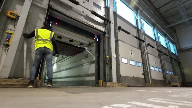 A low angle shot on a warehouse floor as a worker wearing a high visibility vest picks up a long metal load bar and walks into an empty semi trailer at a loading dock