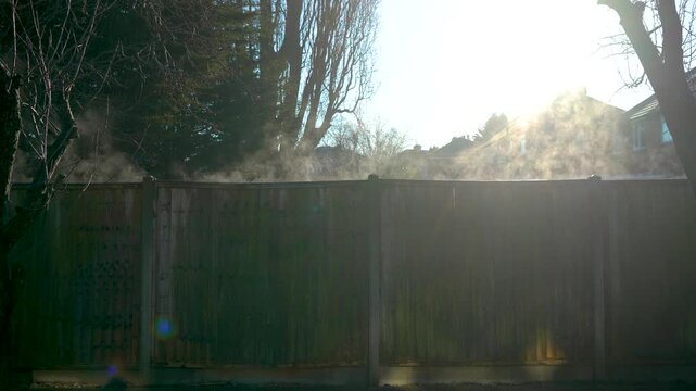Residential area fire emergency unfolding in private sector neighborhood. Thick smoke rising above wooden fence and drifting through suburban houses. Accident scene escalating while sunlight