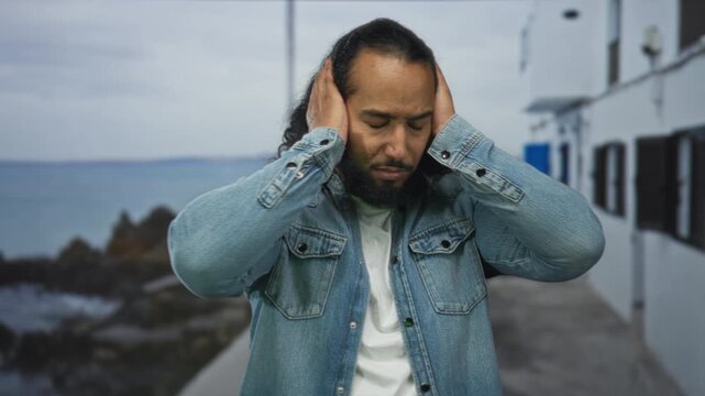 Man in denim jacket holds hands to ears beside rocky seaside promenade on street with white buildings and pier; frustration isolation.