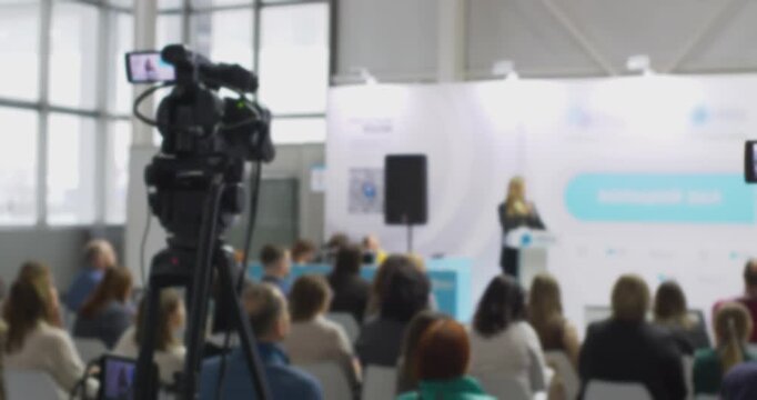 Professional video camera on a tripod recording a female speaker giving a presentation at a business forum, with a blurred audience listening attentively in the background