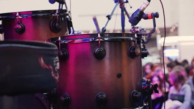 Professional drum kit and vocal microphones set up on an empty stage in a mall atrium. Close-up of a drum set and microphone stands on a vacant concert platform. Excited crowd of young fans.