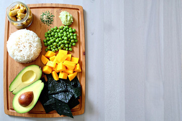 Vibrant healthy food platter featuring white rice, green peas, diced mango, ripe avocado, nori seaweed on wooden cutting board in modern professional food photography