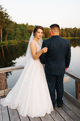 Naklejka premium Romantic wedding couple embracing on wooden pier by calm lake. Bride in elegant white dress and veil looks lovingly at groom in suit. Tender moment, soft natural light, intimate atmosphere.