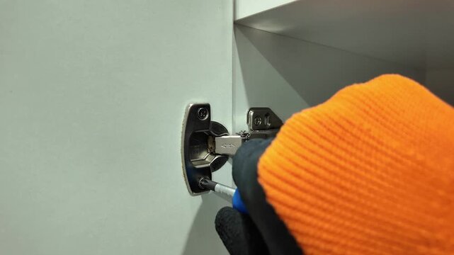 A gloved worker installs a cabinet hinge with a screwdriver during furniture assembly