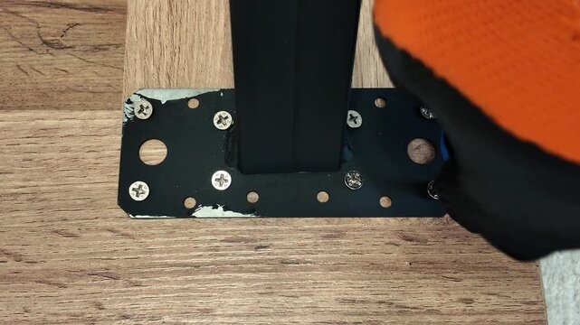 Close-up of a mechanic wearing protective gloves using a screwdriver to screw a screw into a metal plate on a wooden table