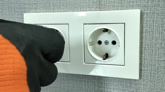 A professional electrician wearing protective gloves uses a screwdriver to install a new double white outlet into a wall