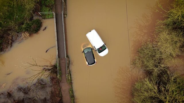 Flood disaster submerging cars in deep muddy water on rural roadway. Heavy rain aftermath leaving vehicles stranded near small bridge and flooded countryside path. Extreme weather crisis causing