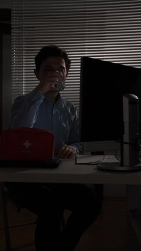 Vertical video overworked office worker taking pills with a glass of water at his desk late at night, stressed and battling burnout