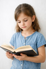 Thoughtful little girl reading book while standing near white wall, calm childhood education and learning concept portrait.