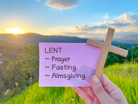 Purple note with Lent, prayer, fasting, and almsgiving text held with wooden cross against sunset landscape, symbolizing the pillars of Lent season.