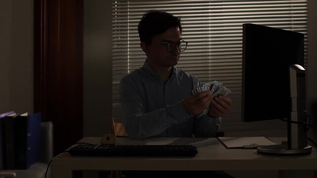 Man in an office at night carefully counting a stack of hundred dollar bills, representing income, financial success, profit, greed, or illicit earnings from his busy work schedule
