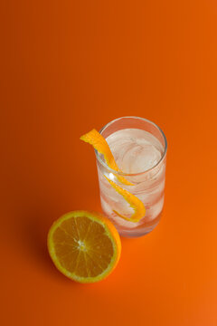An overhead view of an iced coktail in a Hi Ball Galss with an orange twist and half an orange against an orange background