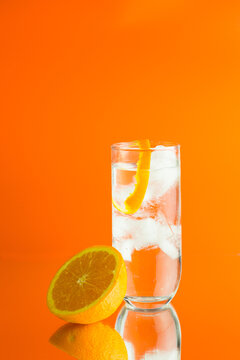 An iced cocktail in a HiBall galss with a twist of orange peel and half an orange against an orange background with a reflection of the orange and the glass