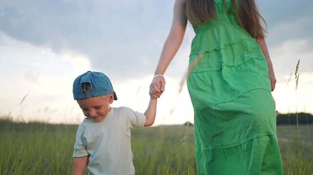 Mother holds son hand walking through meadow field at sunset. Woman with child walks in grass. Family enjoys meadow nature. Boy and mother in field. Sunset walk through tall grass together.