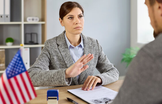 Immigration denial and rejection by consulate officer showing refusal gesture to applicant during visa interview. Female US visa lawyer and man sitting at desk with United States of America flag