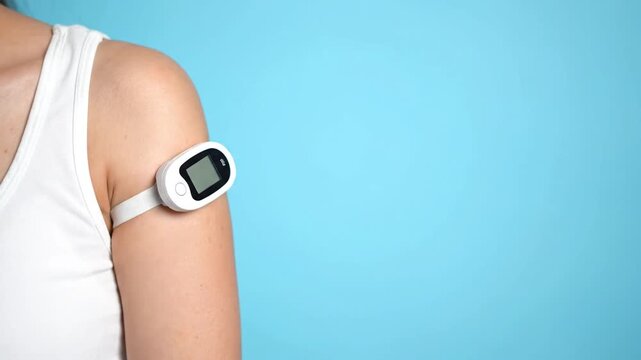 The video shows a close-up of a person's arm wearing a white health monitor, possibly a continuous glucose monitor (CGM), on a light blue background. The device's screen is visible, hinting at contin