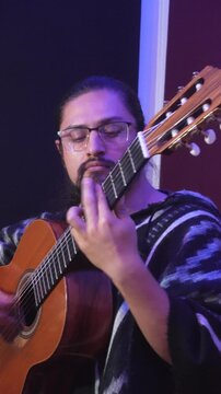 Vertical medium shot of a male guitarist with glasses and a beard, performing with deep concentration, wearing a traditional black and white patterned poncho.