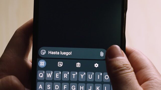 Close-up of hands holding a smartphone, typing the Spanish farewell "Hasta luego!" on a dark-themed digital keyboard. Concept of communication, social media, and digital connection.