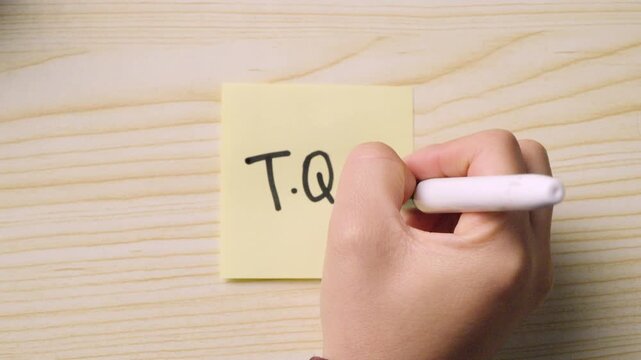 Close-up of a hand using a black marker to write the Spanish acronym "T.Q.M" (Te Quiero Mucho) and a heart on a yellow sticky note. Concept of friendship and love.