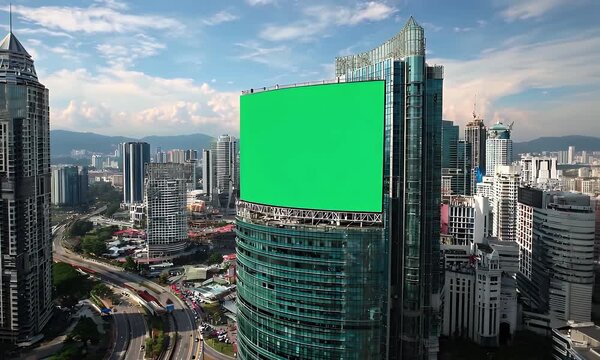 Aerial view of a modern cityscape with a large green billboard on a skyscraper amidst high-rise buildings and busy streets under a partly cloudy blue sky.