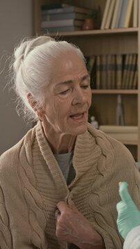 Vertical shot of brunette nurse with rubber gloves checking throat of senior lady with help of tongue depressor and medical penlight