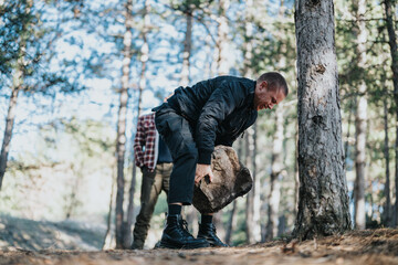 A man in dark clothing lifts a heavy rock on a forest trail. Another person stands in the background, among trees and sunlight.