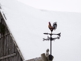 Traditional metal wind vane with a rooster silhouette above a rustic wooden building under heavy winter snow. Rooster weather vane on a snow-covered rooftop in Vasknarva, during a quiet winter day.  © Kaja