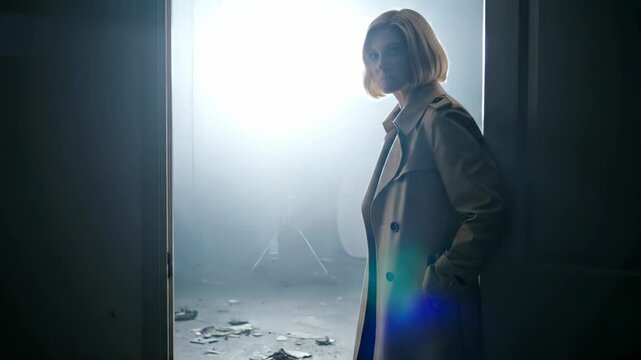 Woman stands in doorway into light. Portrait shows silhouette in trenchcoat. Hands in pocket convey calm. Backlight creates fog and lensflare. Empty room and scattered debris add mystery. Moody tone.