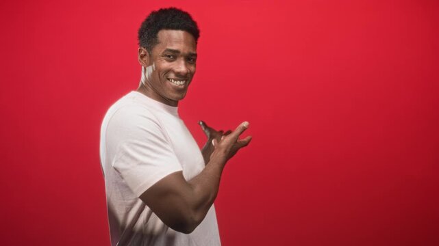 Man pointing fingers with visible hand and smiling, wearing white tshirt in studio red backdrop; confidence charm.