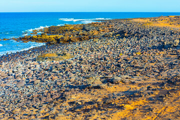 Shoreline blends dark rock volcanic formations with sandy patches, while gentle waves roll. Rugged textures with bright coastal serenity