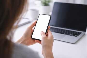 Woman holding smartphone with blank screen in front of laptop on white desk in bright office environment JPG image