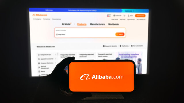 Alibaba app running on a modern mobile phone. The smartphone is being held in landscape mode, in the darkness. A PC monitor with the Alibaba.com website in the background.